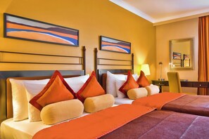 A hotel room with two beds, each with orange bedding and pillows, a desk with a lamp, and two framed abstract paintings on the wall. - Corinthia St George's Bay (St. Julian's)