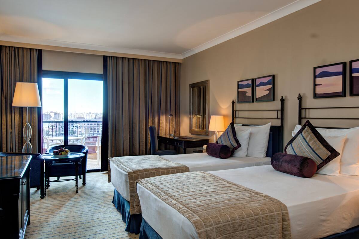Ein Hotelzimmer mit zwei Betten, einem Schreibtisch und einem Stuhl. Ein großes Fenster mit Vorhängen bietet Blick auf die Stadt, und an der Wand hängt ein Kunstwerk.