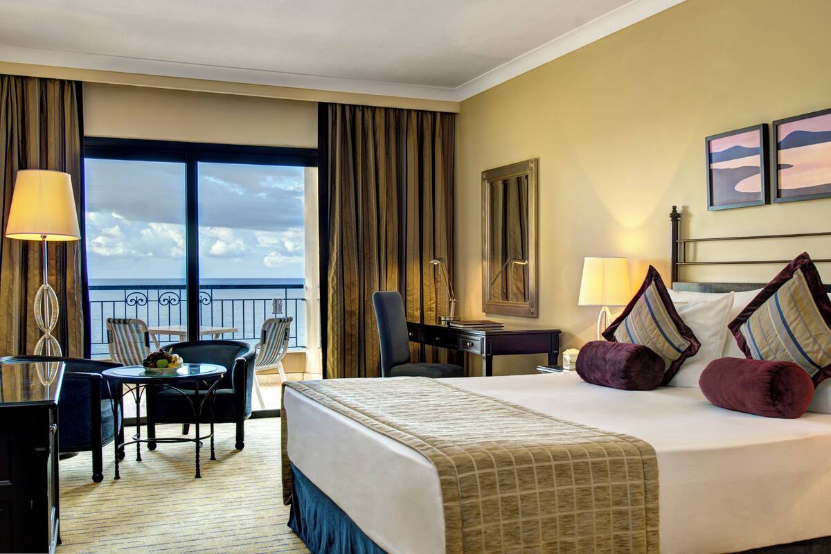 Ein Hotelzimmer mit Bett, Schreibtisch, Stuhl, kleinem Tisch mit Lampe und Blick auf den Himmel durch große Fenster.