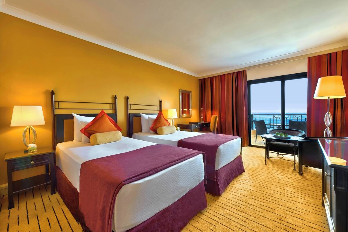 Ein Hotelzimmer mit zwei Betten, einem Schreibtisch, einem Stuhl, einer Lampe und Blick nach draußen.