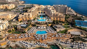 An aerial view of a coastal hotel with multiple buildings, a central swimming pool, and a tennis court. - Corinthia St George's Bay (St. Julian's)