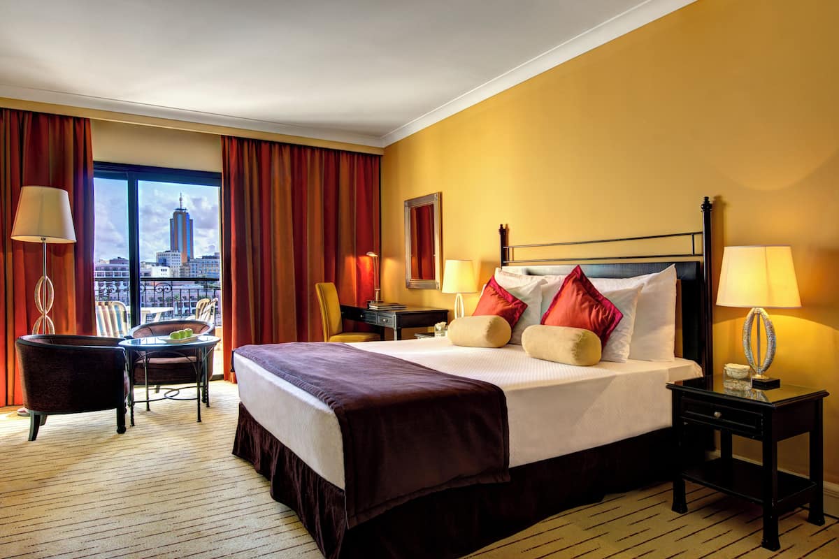 Ein Hotelzimmer mit einem großen Bett, zwei Nachttischen mit Lampen, einem Sessel, einem kleinen Tisch mit einem Glas und Blick auf eine Stadtkulisse durch ein großes Fenster.