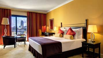A hotel room with a large bed, two bedside tables with lamps, a chair, a small table with a glass, and a view of a cityscape through a large window.