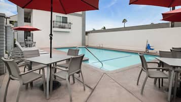 An outdoor pool area with grey lounge chairs and tables, red umbrellas, and a clear blue sky.