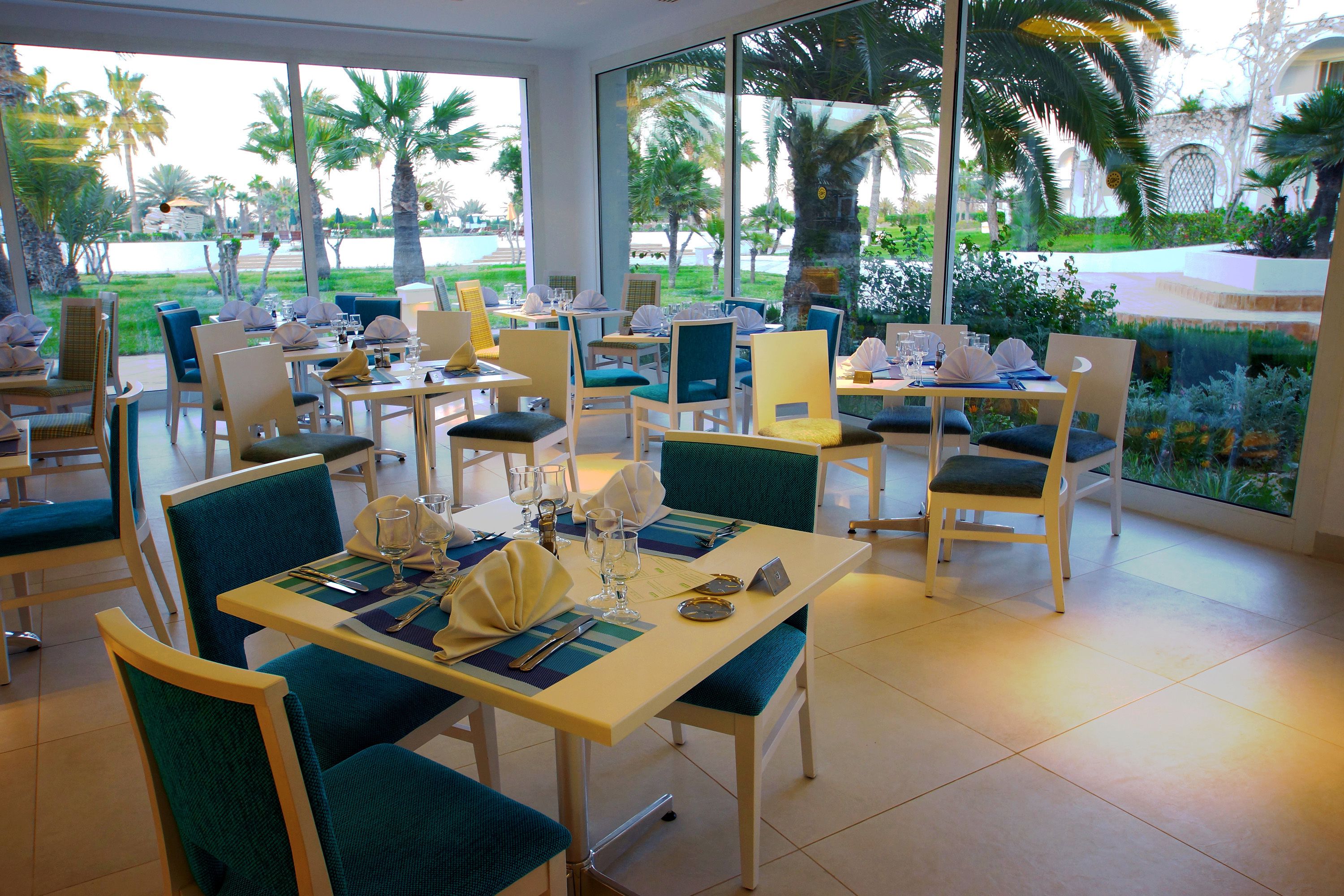 Un restaurante con mesas dispuestas para comer, sillas y vistas a palmeras y una piscina.