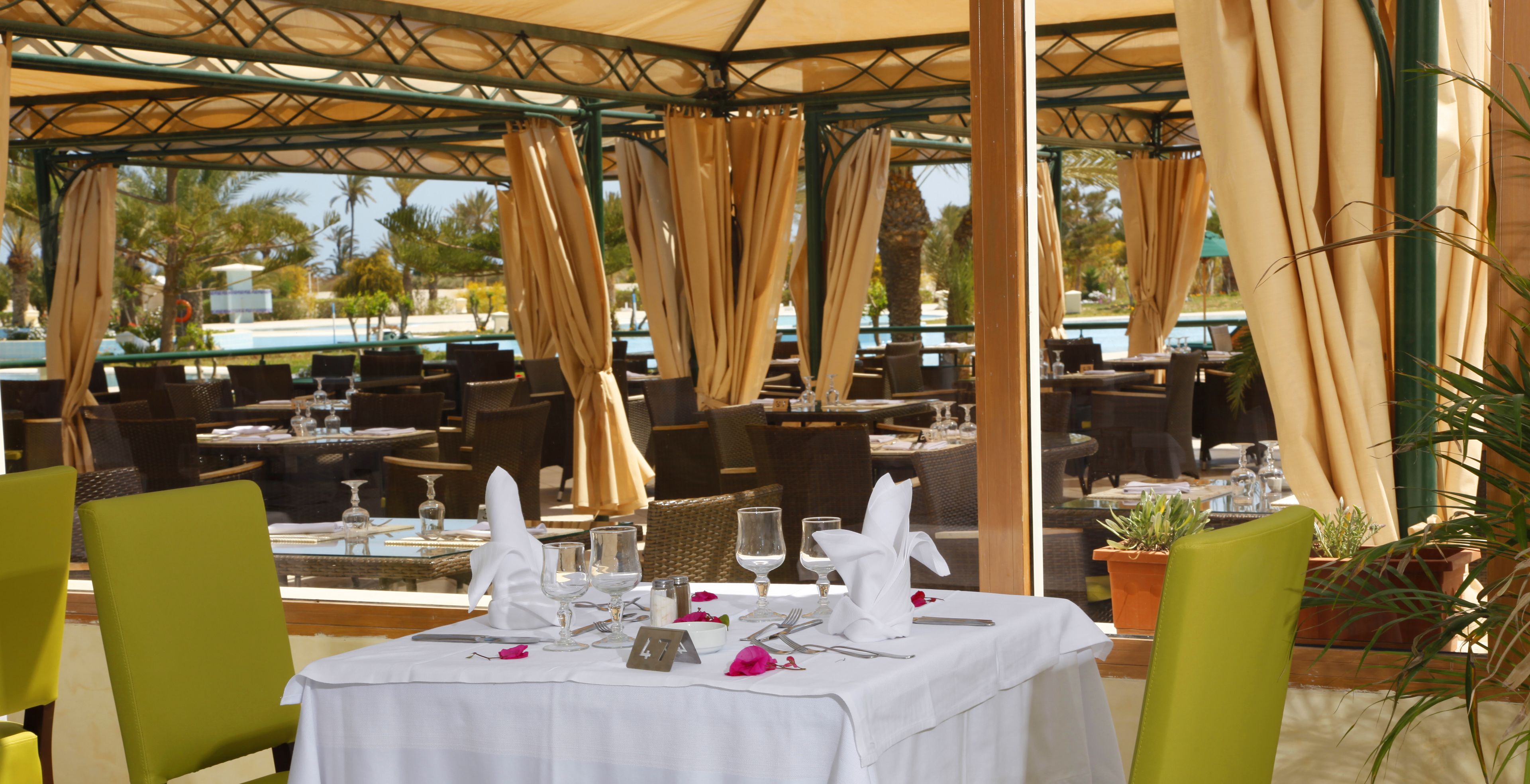 An outdoor dining area with tables set for two, covered by a pergola with beige curtains.