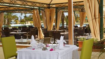 An outdoor dining area with tables set for two, covered by a pergola with beige curtains.
