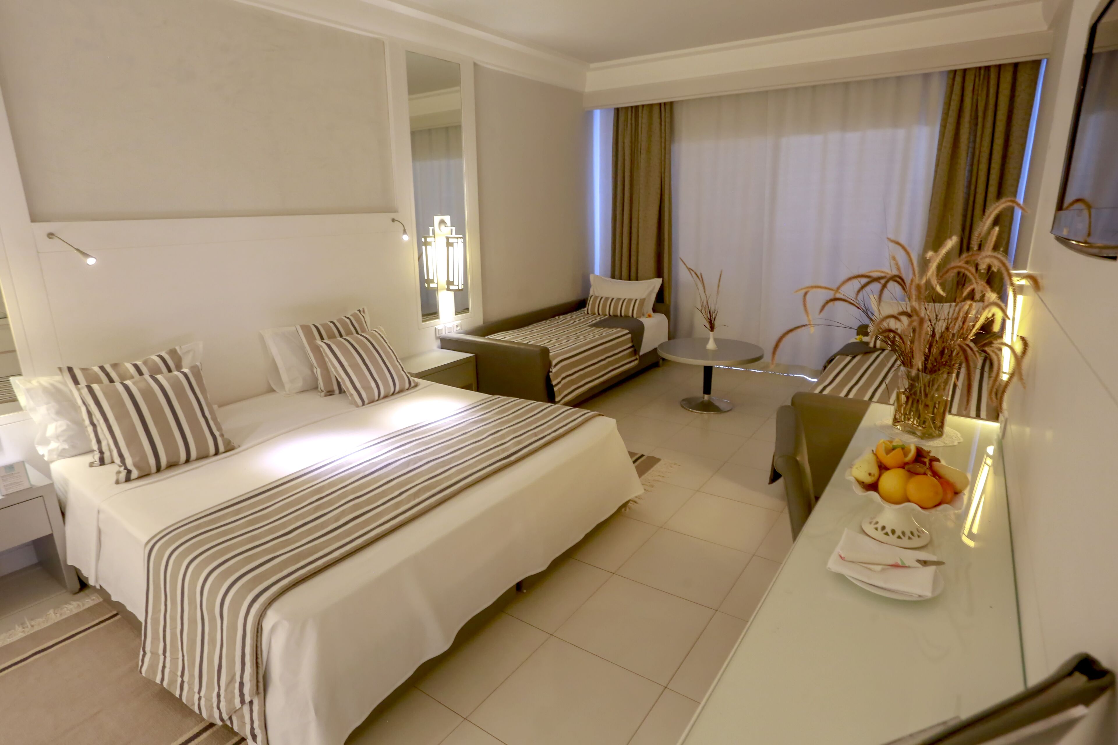 Una habitación de hotel con una cama grande, una cama más pequeña, un escritorio con un florero y una mesa de comedor con fruta.