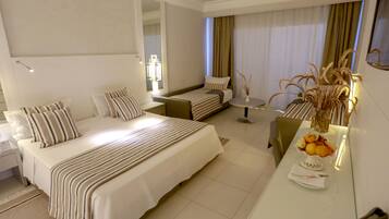 Una habitación de hotel con una cama grande, una cama más pequeña, un escritorio con un florero y una mesa de comedor con fruta.