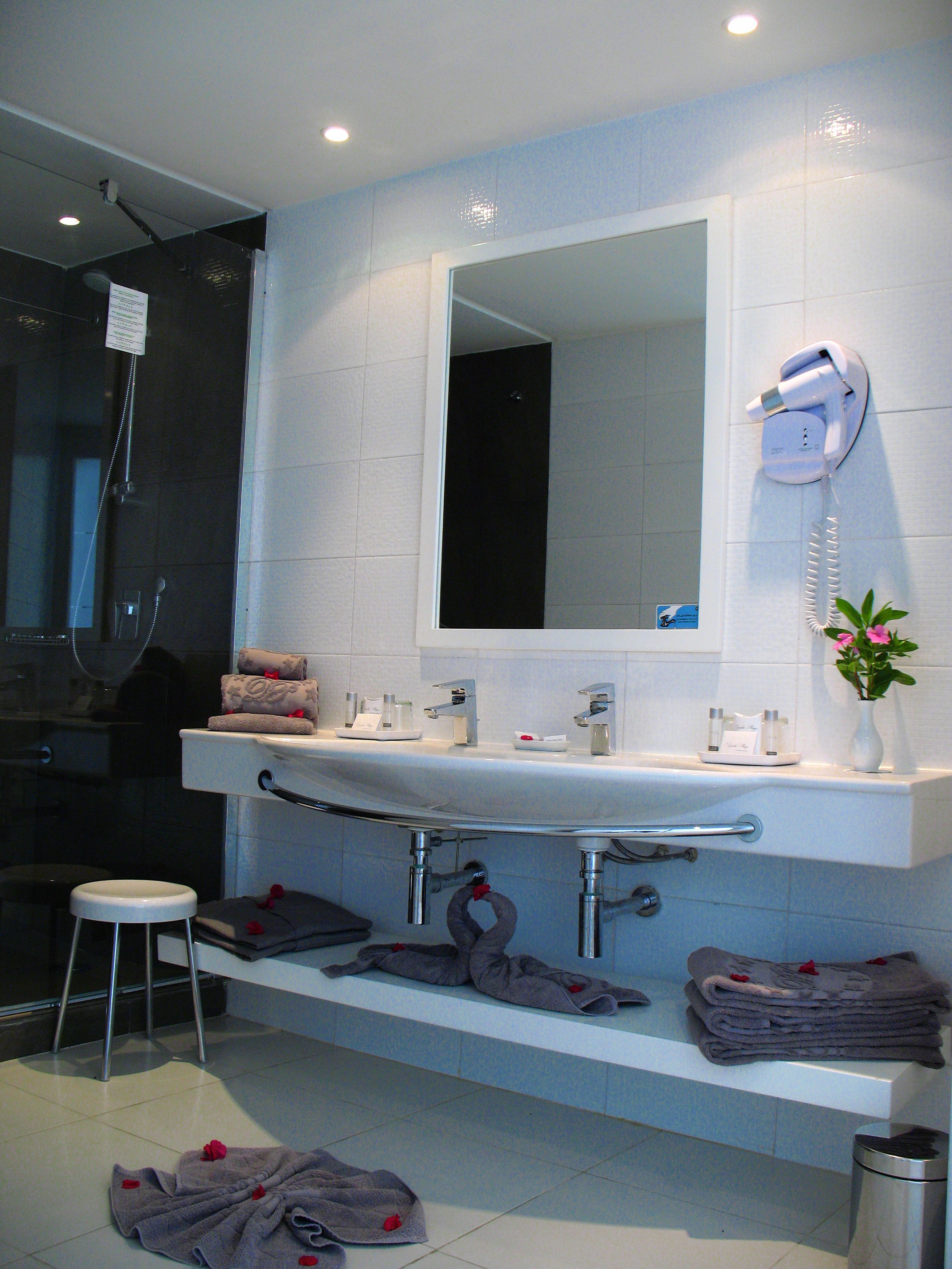 Un baño moderno con un lavabo blanco, espejo y un secador de pelo montado en la pared. Hay un perchero con toallas dobladas y un taburete pequeño.
