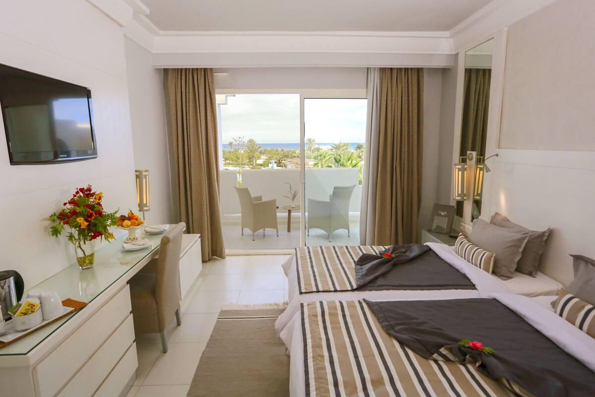 A hotel room with two beds, a desk, a TV, and a balcony with a view.