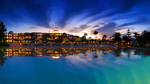 A hotel with a pool reflecting the building and palm trees, surrounded by umbrellas and lounge chairs. - Djerba Plaza Thalasso & Spa (Djerba Midun)