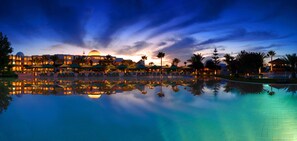 A hotel with a pool reflecting the building and palm trees, surrounded by umbrellas and lounge chairs. - Djerba Plaza Thalasso & Spa (Djerba Midun)