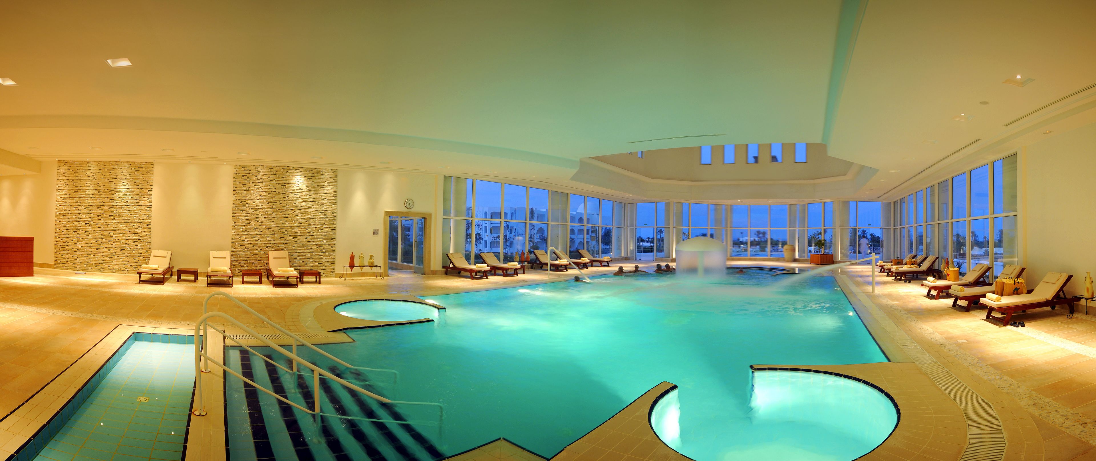 An indoor swimming pool with multiple lounge areas and a stone wall.