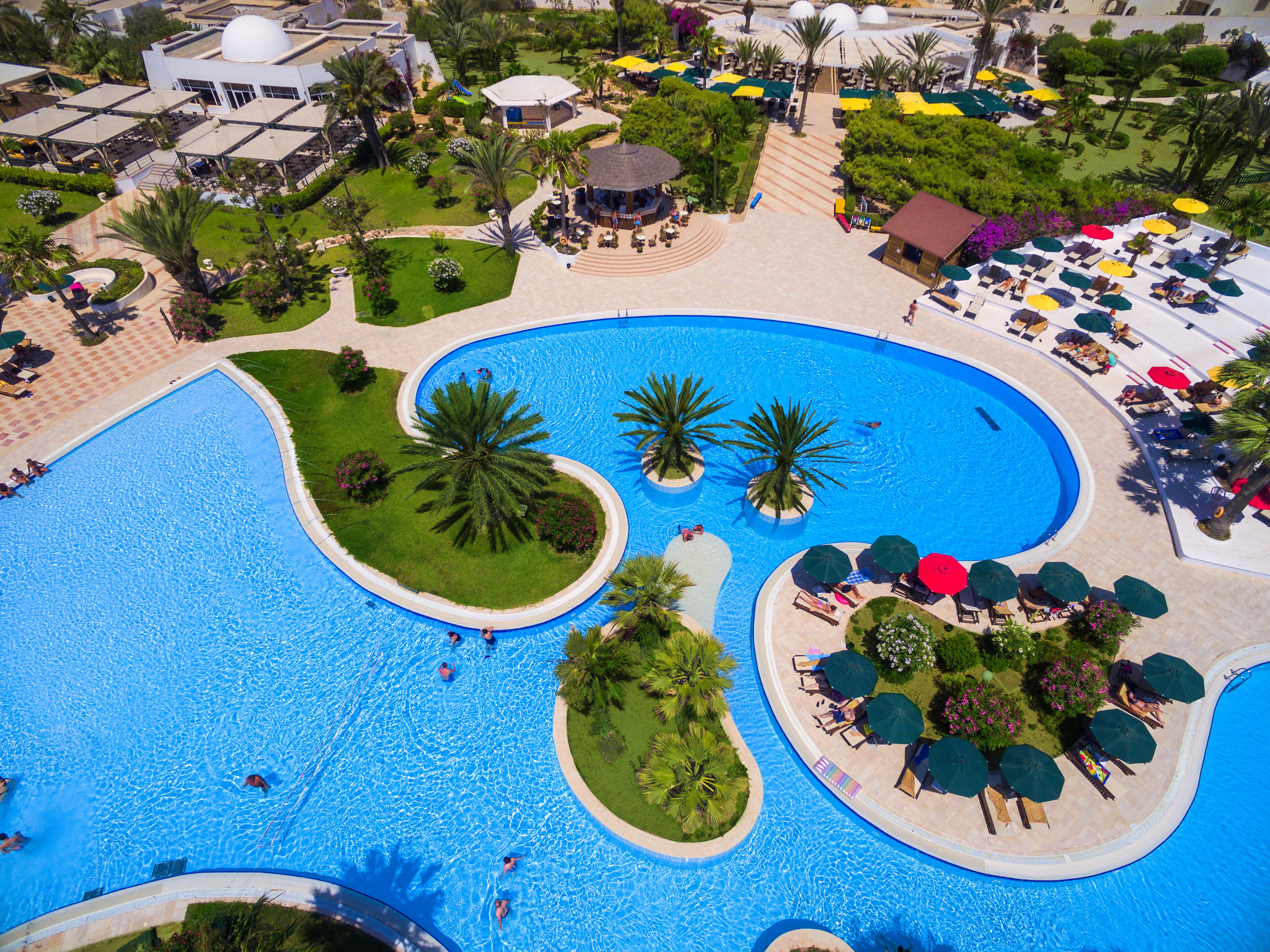 Vista aérea de um resort com uma piscina central ampla, rodeada por palmeiras e espreguiçadeiras.