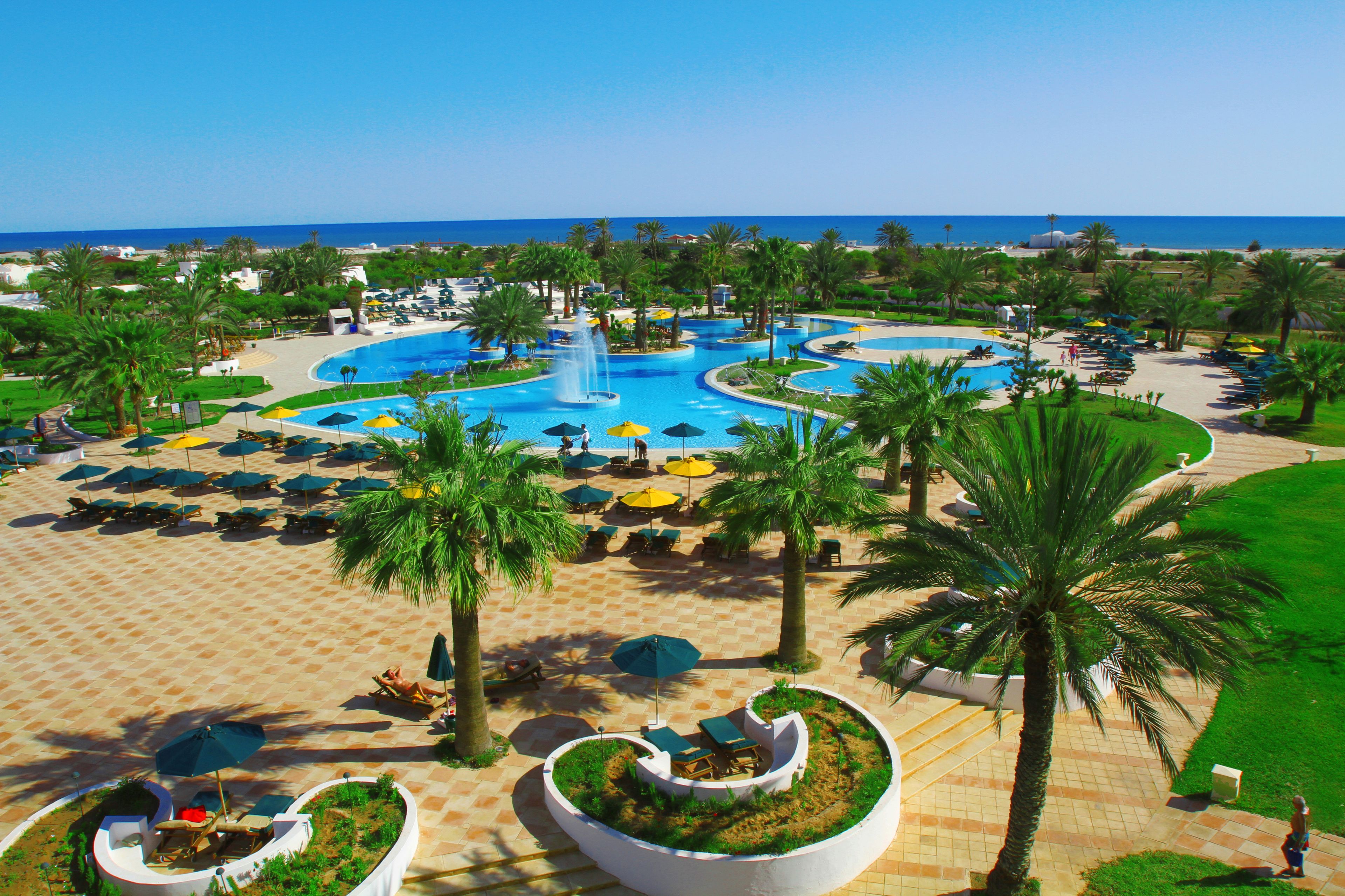 A resort with a central swimming pool, sun loungers, palm trees, and a view of the ocean.