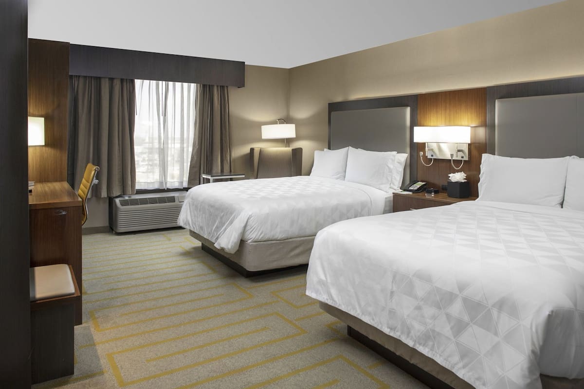 A hotel room with two beds, a desk, a chair, a lamp, and a window with curtains.