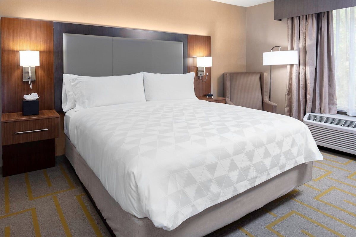 A hotel room with a large bed, two bedside tables, a chair, a desk, a lamp, and a window with curtains.