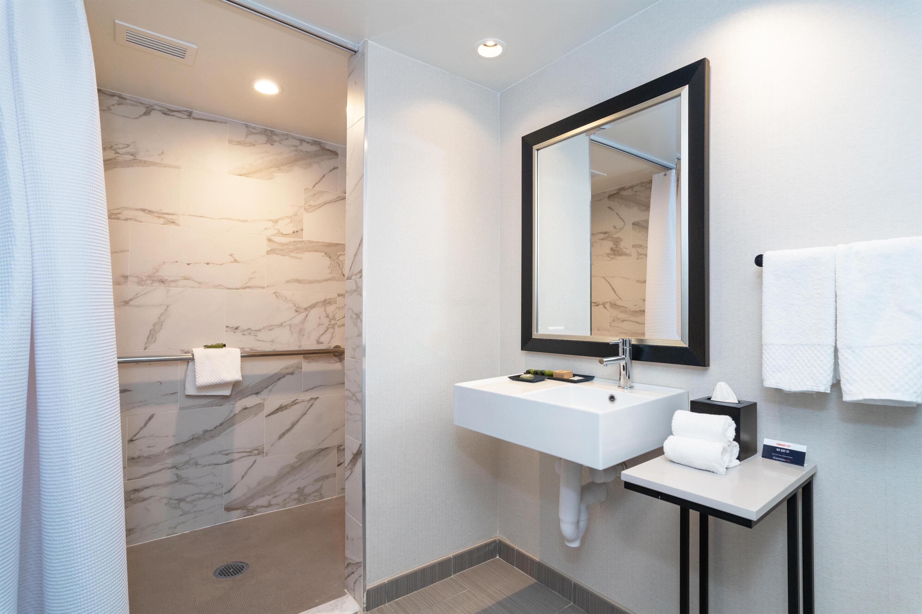A modern bathroom with a marble shower wall, a white sink with a mirror, and a small table with a towel and a tissue box.