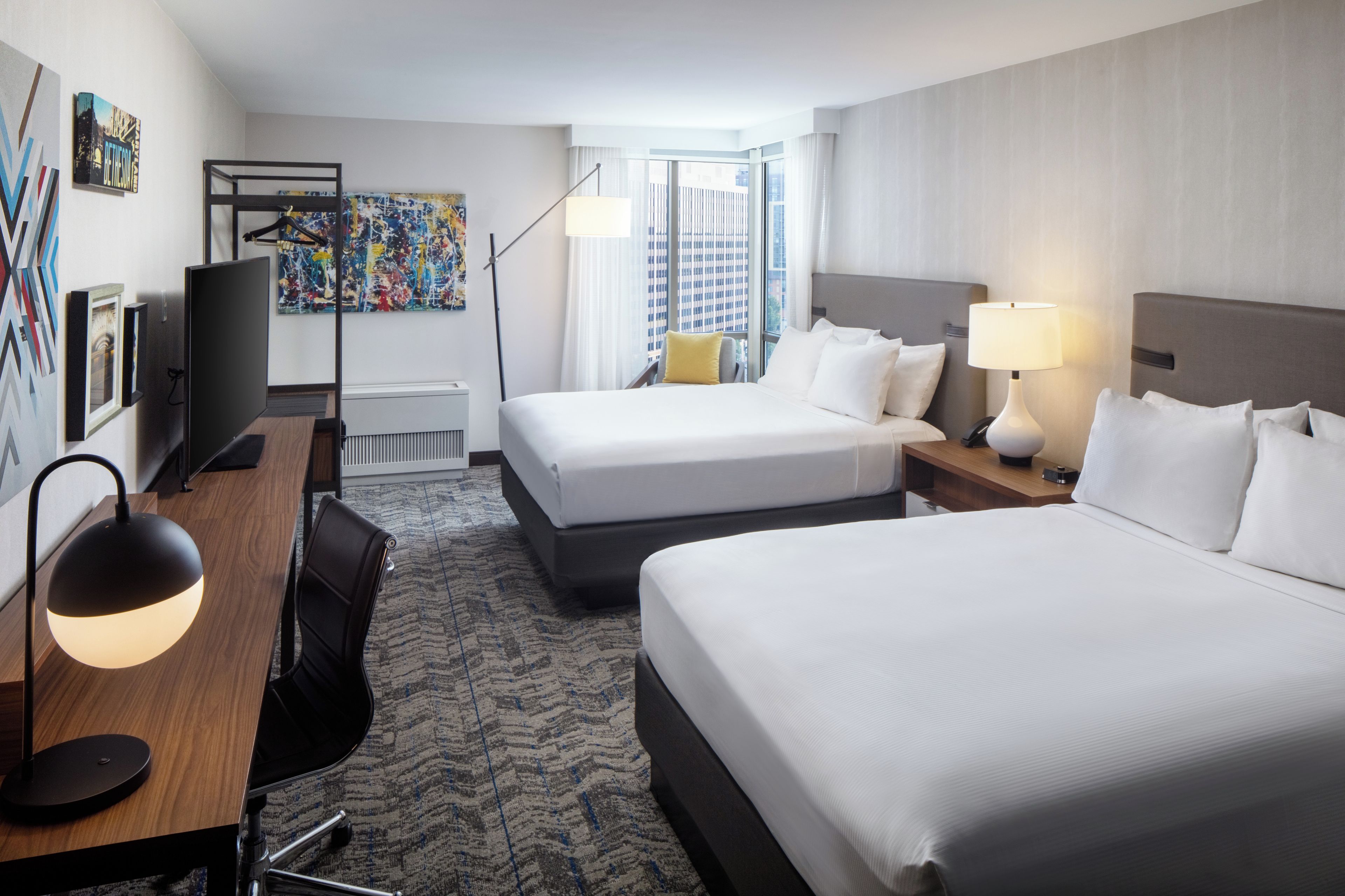 Quarto de hotel com duas camas, uma escrivaninha, uma cadeira, televisão e obras de arte nas paredes.