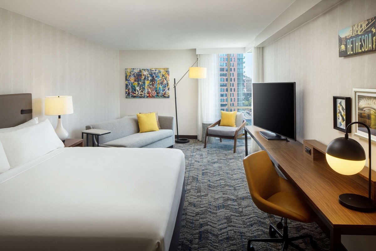 A hotel room with a bed, a desk, a chair, a TV, and a painting on the wall.
