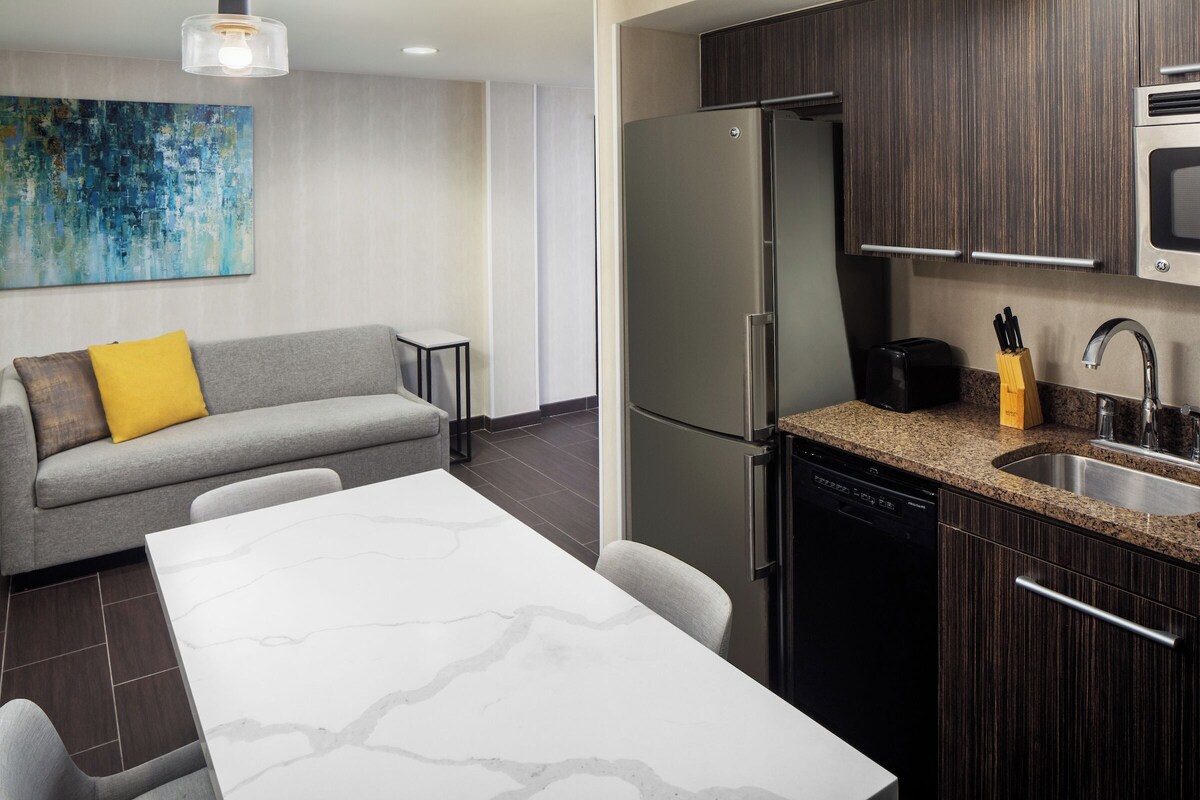 A compact kitchen with a refrigerator, microwave, and dishwasher. There's a gray sofa with a yellow pillow, a white marble table, and a small side table.