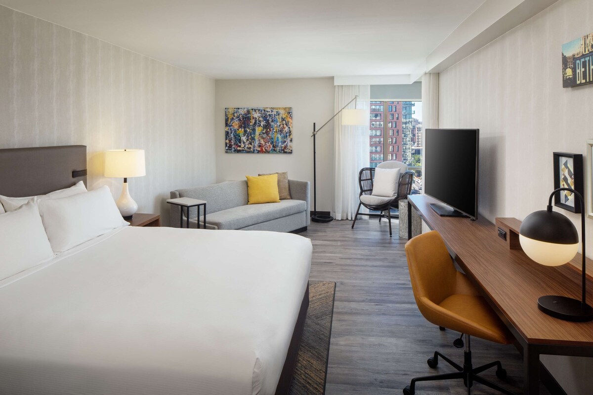 A hotel room with a large bed, a sofa, a desk, a chair, a TV, and a painting on the wall.