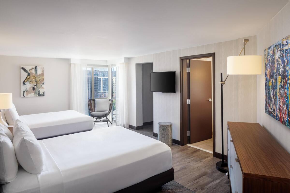 Et hotelværelse med to senge, et fjernsyn, et skrivebord, en stol og et stort vindue.