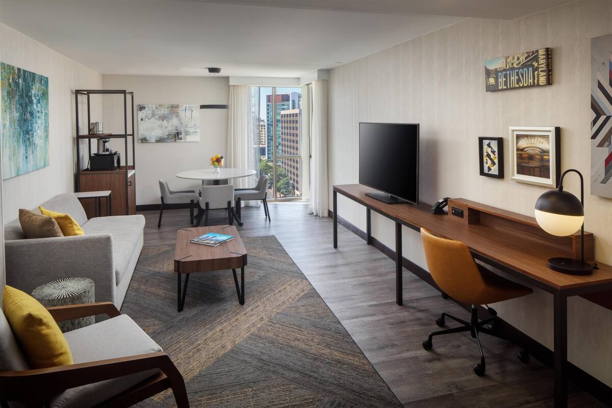 Et moderne hotelværelse med sofa, skrivebord, tv, spiseplads og byudsigt.