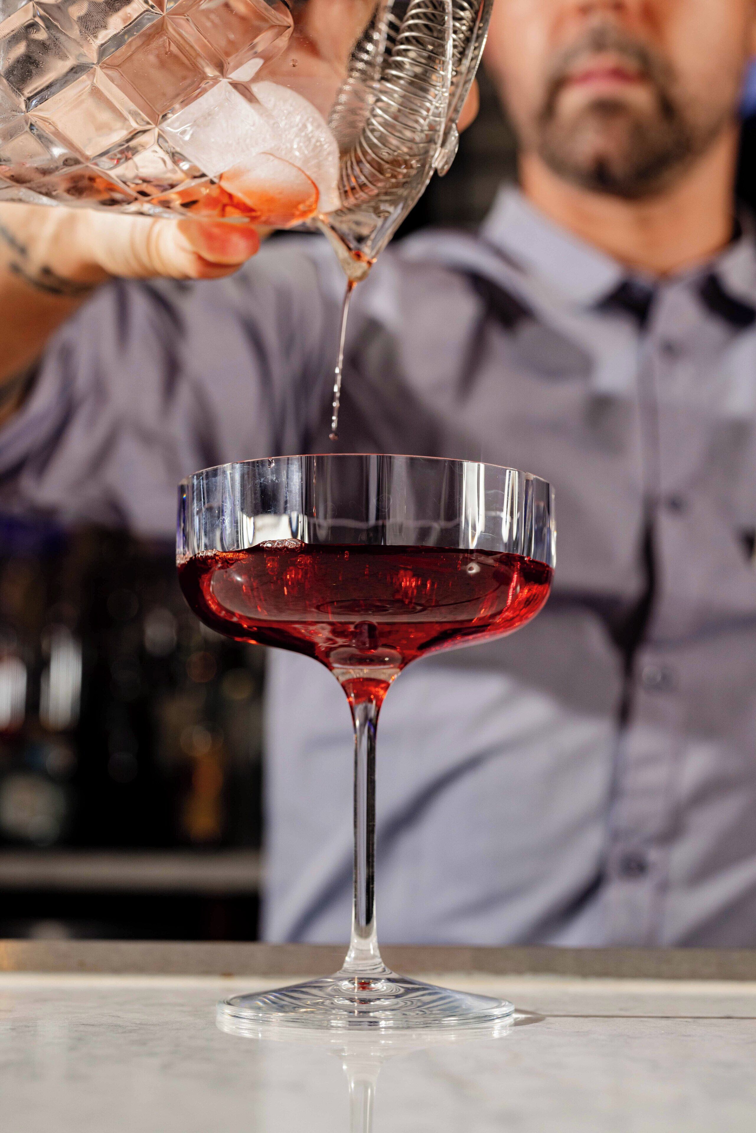 A bartender pouring a red liquid into a stemmed glass.