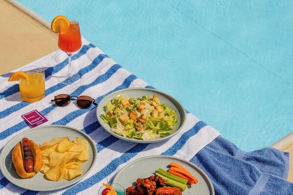 A poolside table with a blue and white striped towel, a plate with a hot dog and chips, a plate with salad and vegetables, a glass of orange juice, and a glass of red drink. - The Bethesdan Hotel, Tapestry Collection by Hilton (Bethesda)