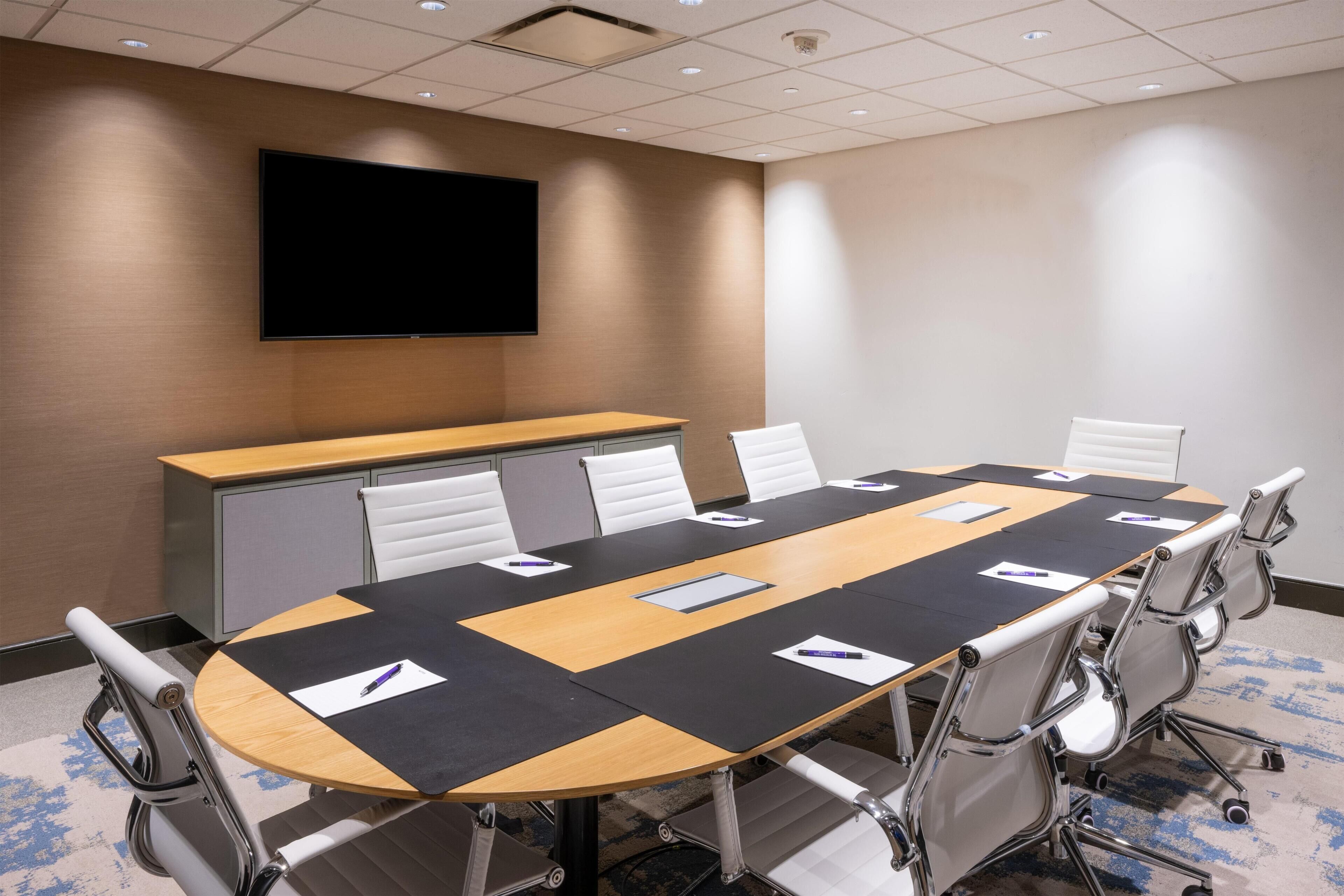 Une salle de conférence avec une longue table, des chaises blanches, un téléviseur à écran plat et un pupitre.