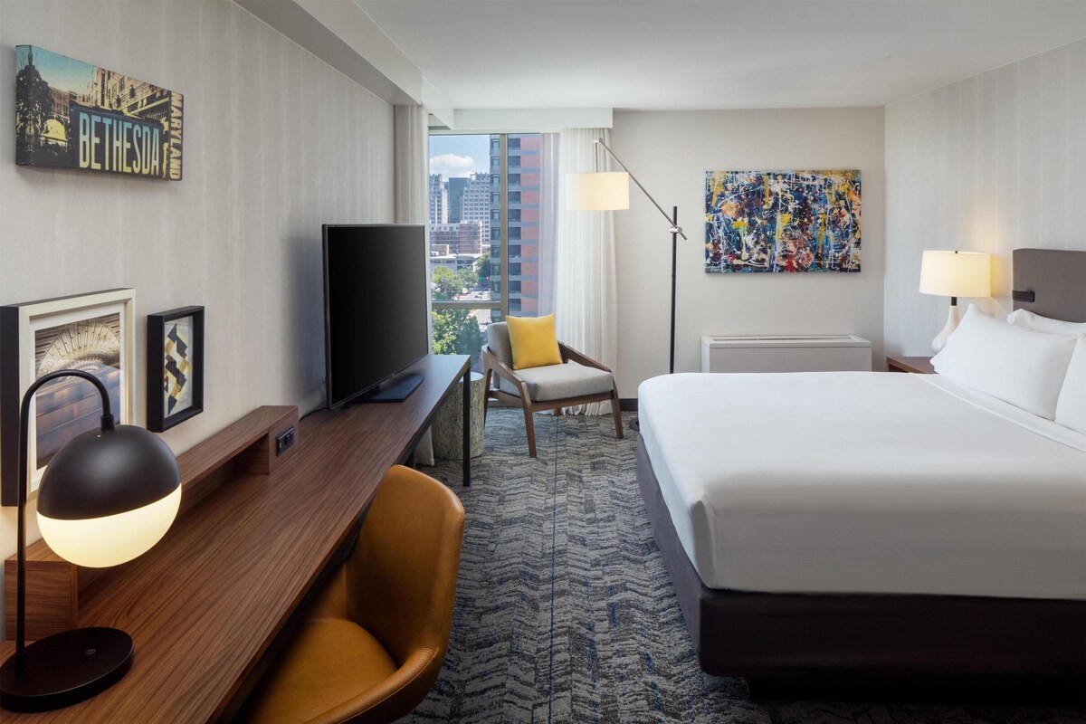 A hotel room with a bed, desk, chair, TV, lamp, and artwork.