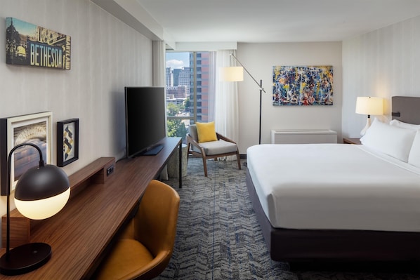 A hotel room with a bed, desk, chair, TV, lamp, and artwork.