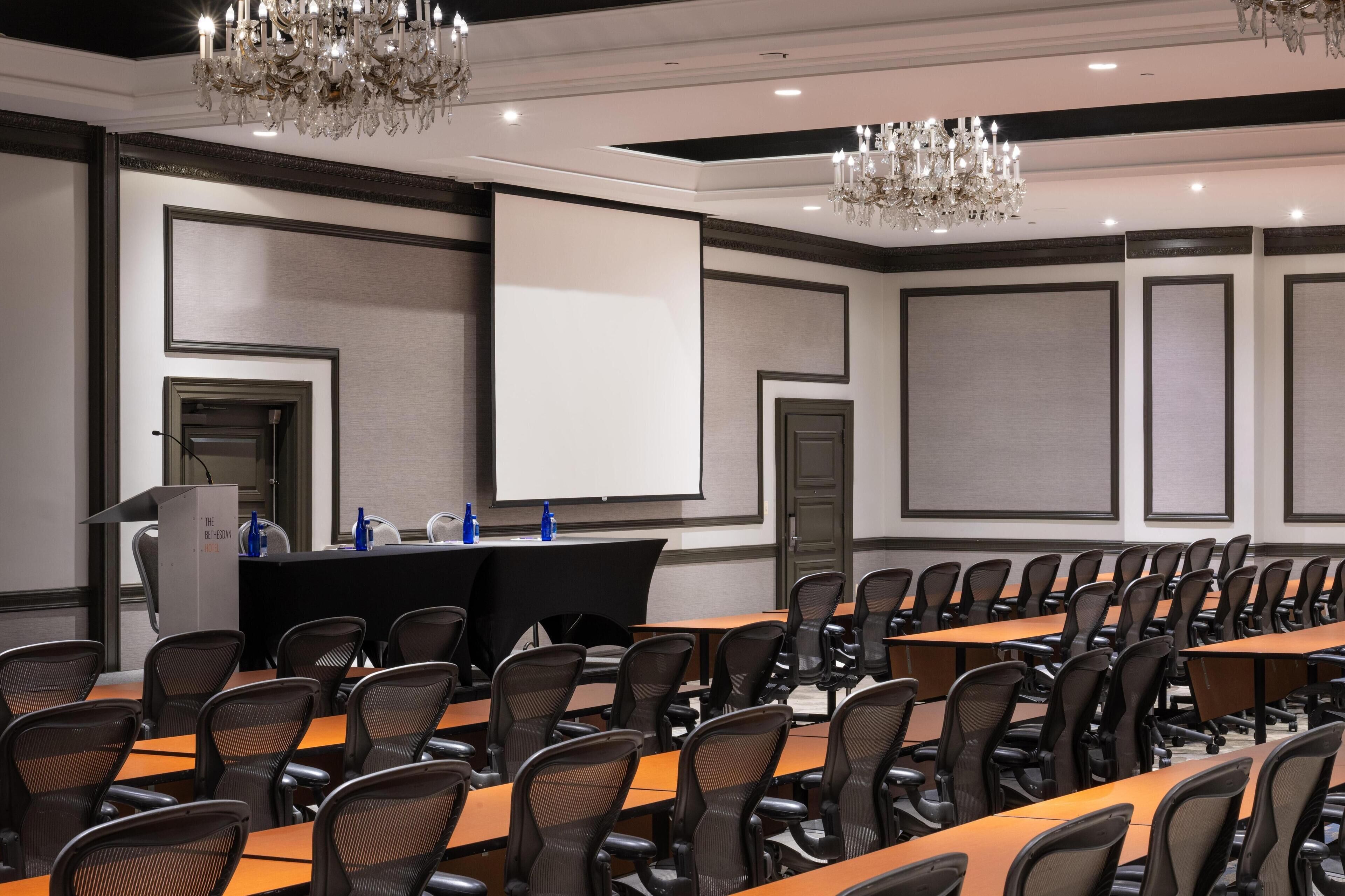 Un salón de conferencias con filas de sillas, un atril, una pantalla y candelabros.