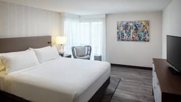 Ein Hotelzimmer mit einem großen Bett, einem Sessel, einem Fernseher, einem Gemälde und einem Fenster mit Vorhängen.