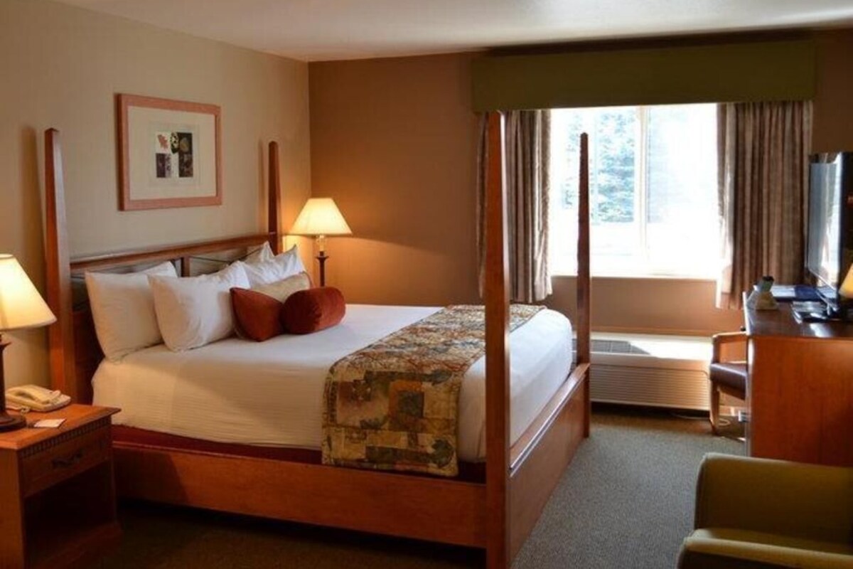 A four-poster bed with a quilt, two bedside lamps, a framed picture on the wall, and a desk with a chair.