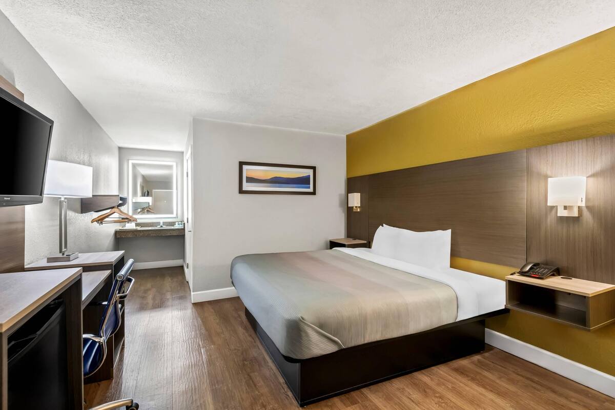 A hotel room with a bed, bedside tables, a desk with a chair, a TV, a mirror, and a painting on the wall.