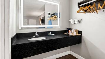 A modern bathroom with a black granite countertop, a white sink, a large mirror with integrated lighting, wooden hangers on a rack, and a towel dispenser.