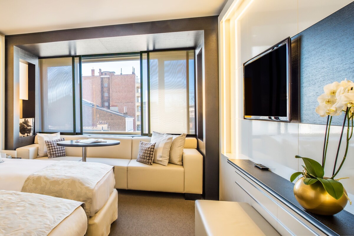 A hotel room with two beds, a sofa, a small table, a TV, and a window with a view of buildings.