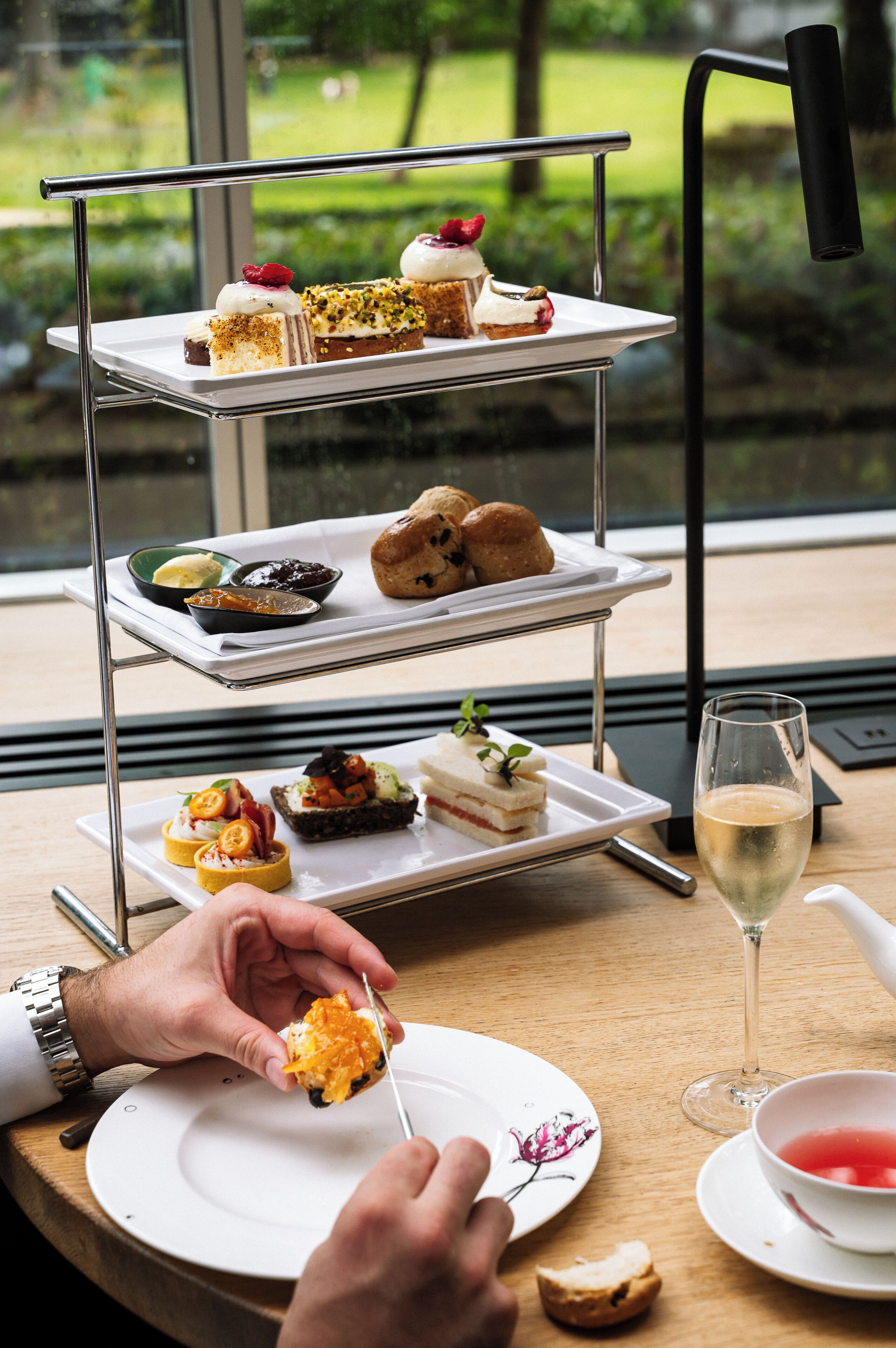 A tiered dessert stand with various pastries and a glass of champagne on a table by a window.