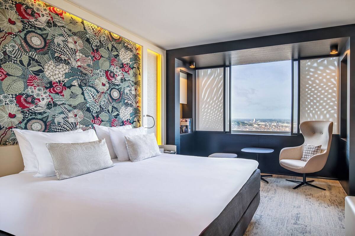 A modern hotel room with a large bed, a seating area with a chair, and a city view through large windows.