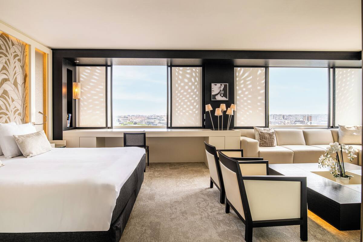 A modern hotel room with a large bed, a sofa, a desk, and a view of the city through large windows.