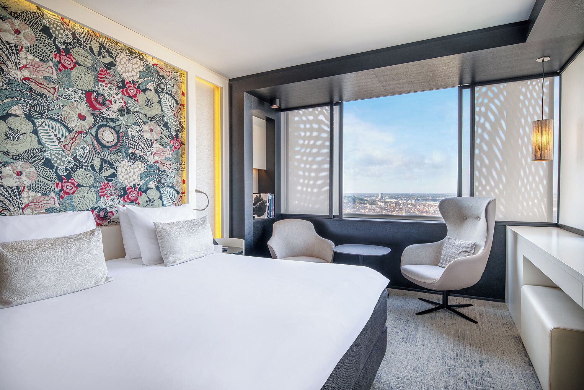 Una habitación de hotel moderna con una cama grande, dos sillones, una mesita y vistas a la ciudad a través de amplios ventanales.