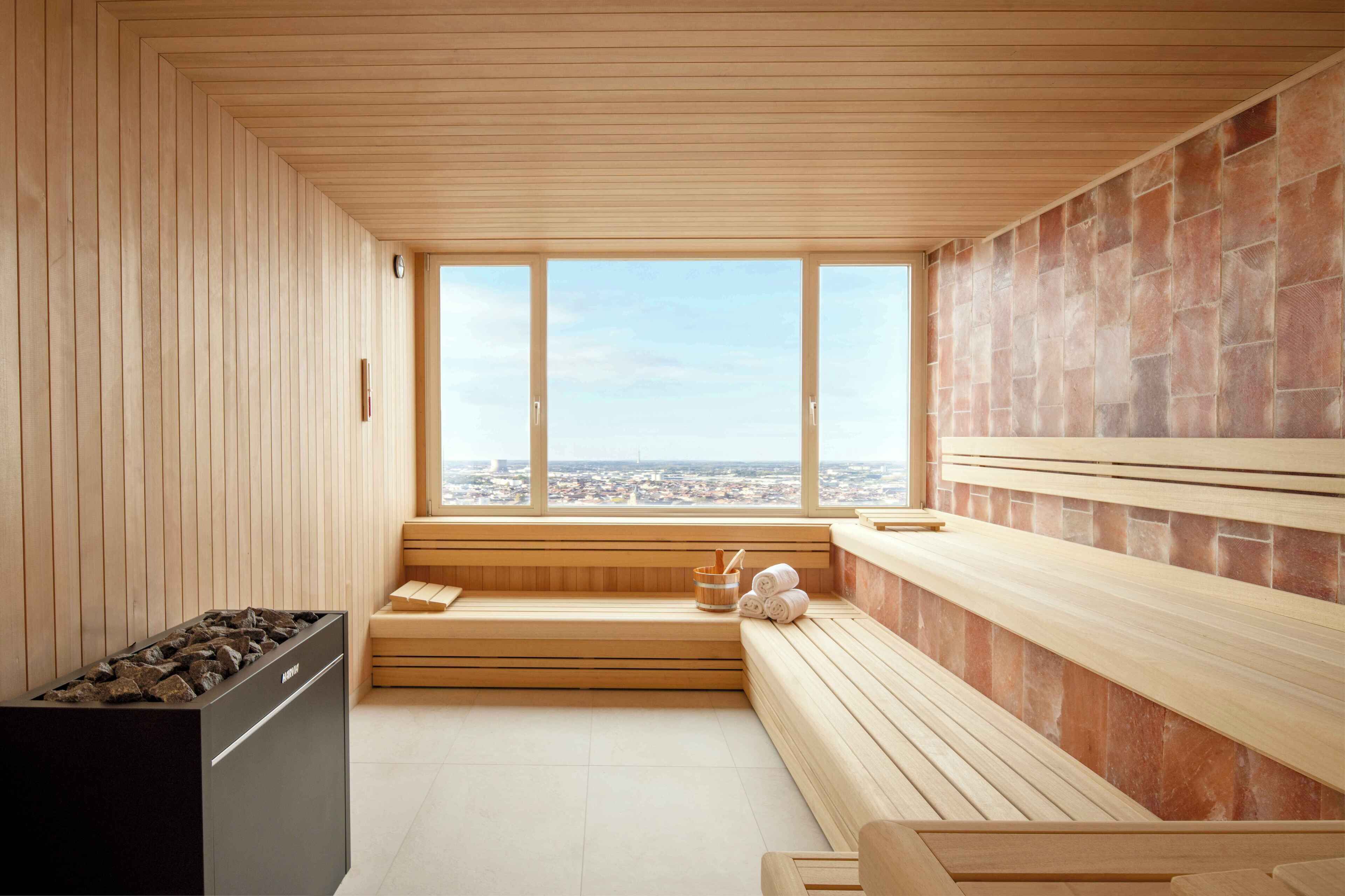 A modern sauna with wooden benches, a stone wall, and a large window offering a city view.
