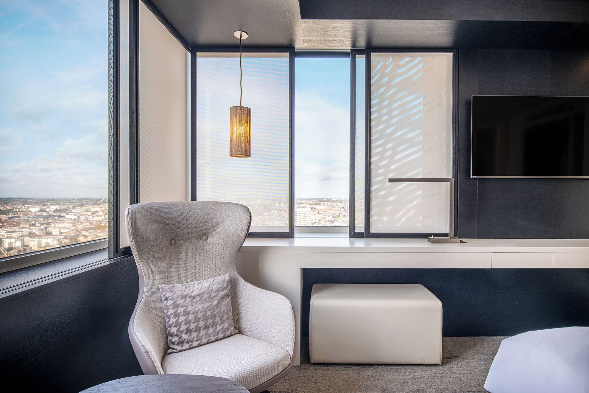 A modern room with a large window, a hanging light fixture, a flat-screen TV, and a cushioned bench.