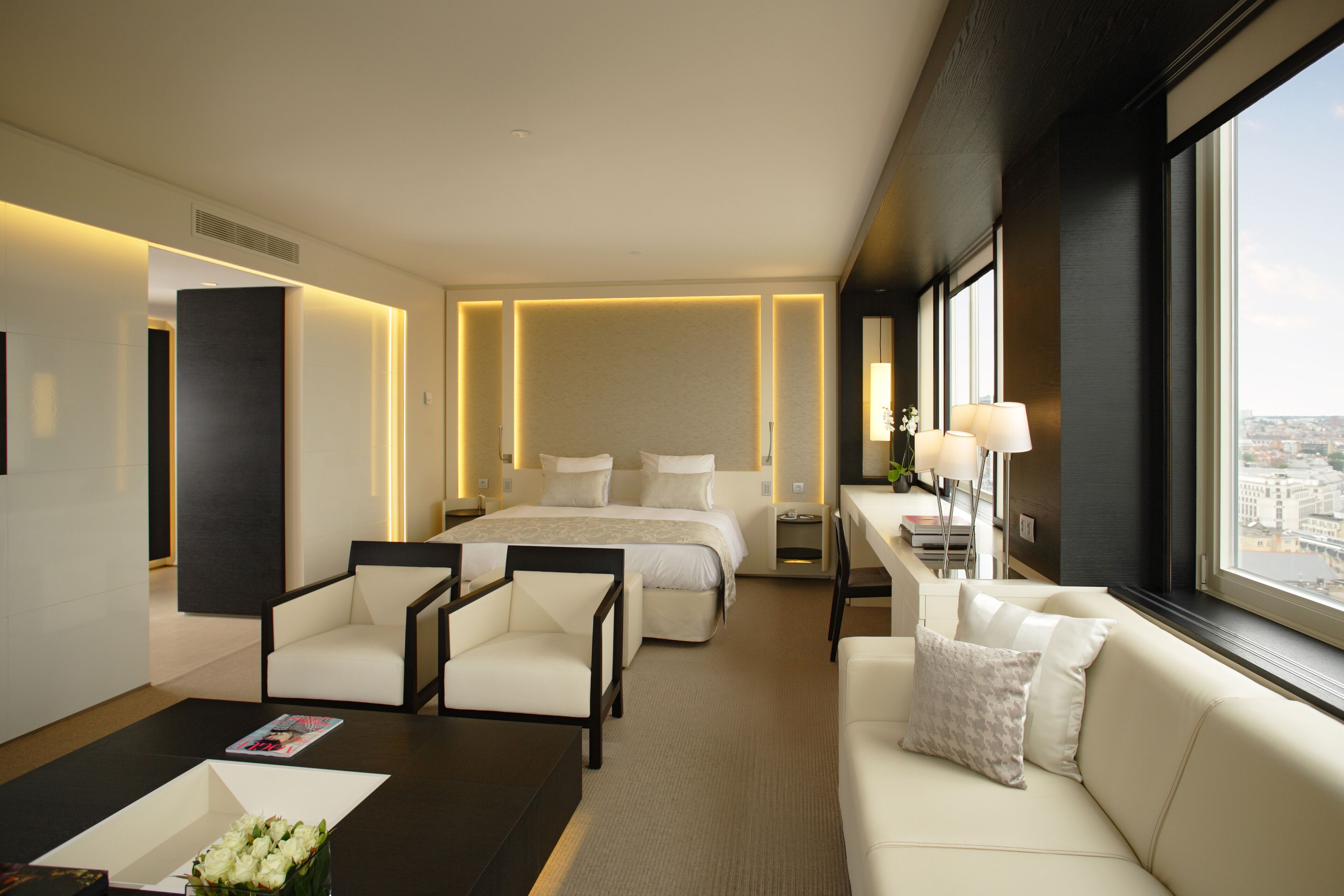 Une chambre d’hôtel moderne, équipée d’un lit, de deux fauteuils blancs, d’un canapé, d’une table basse et offrant une vue sur la ville grâce à de grandes fenêtres.