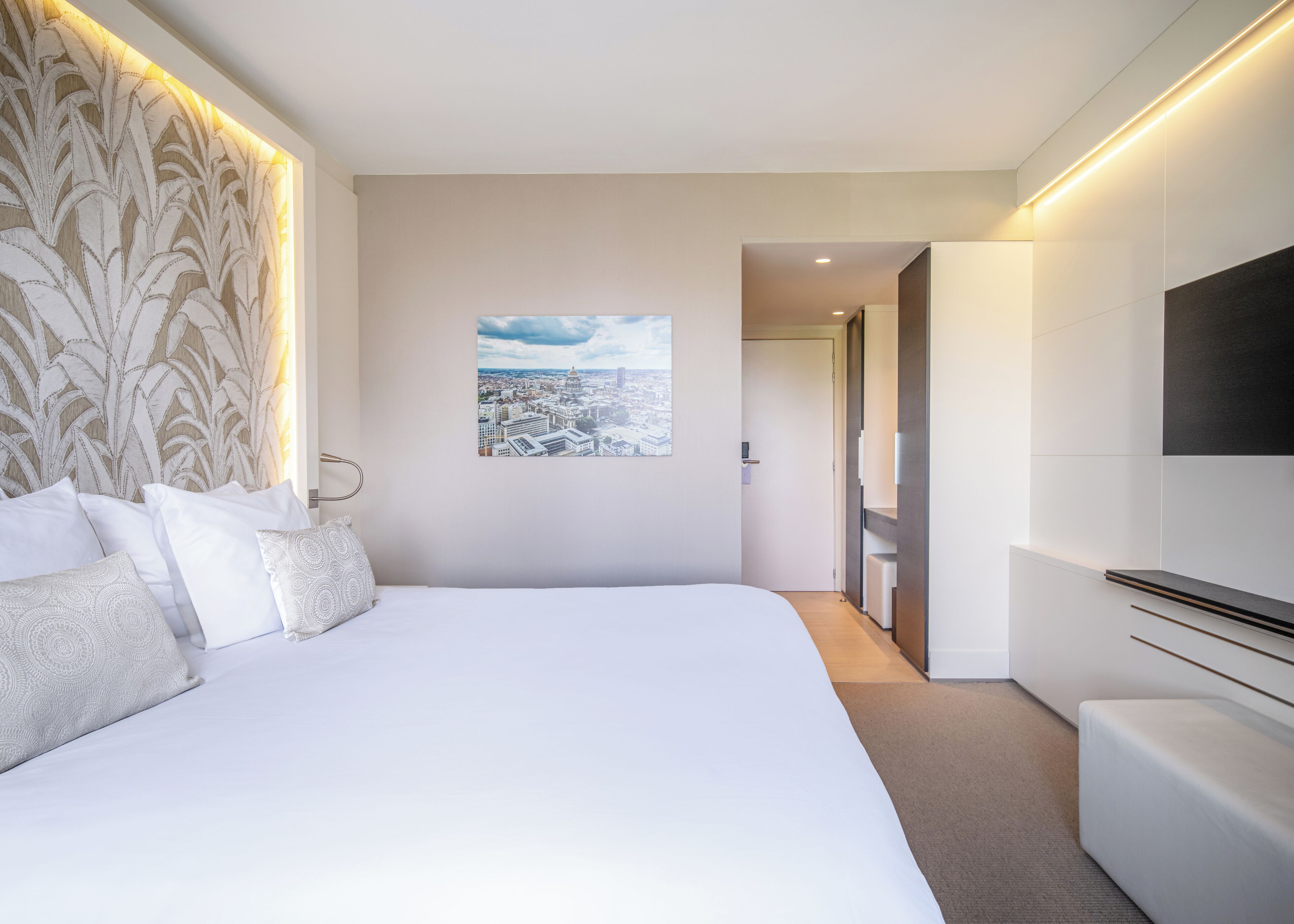 A modern hotel room with a large bed, a cityscape view, a flat-screen TV, and a walk-in closet.