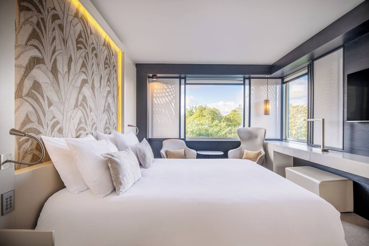 A modern hotel room with a large bed, two armchairs, a small table, and a large window offering a view of greenery.