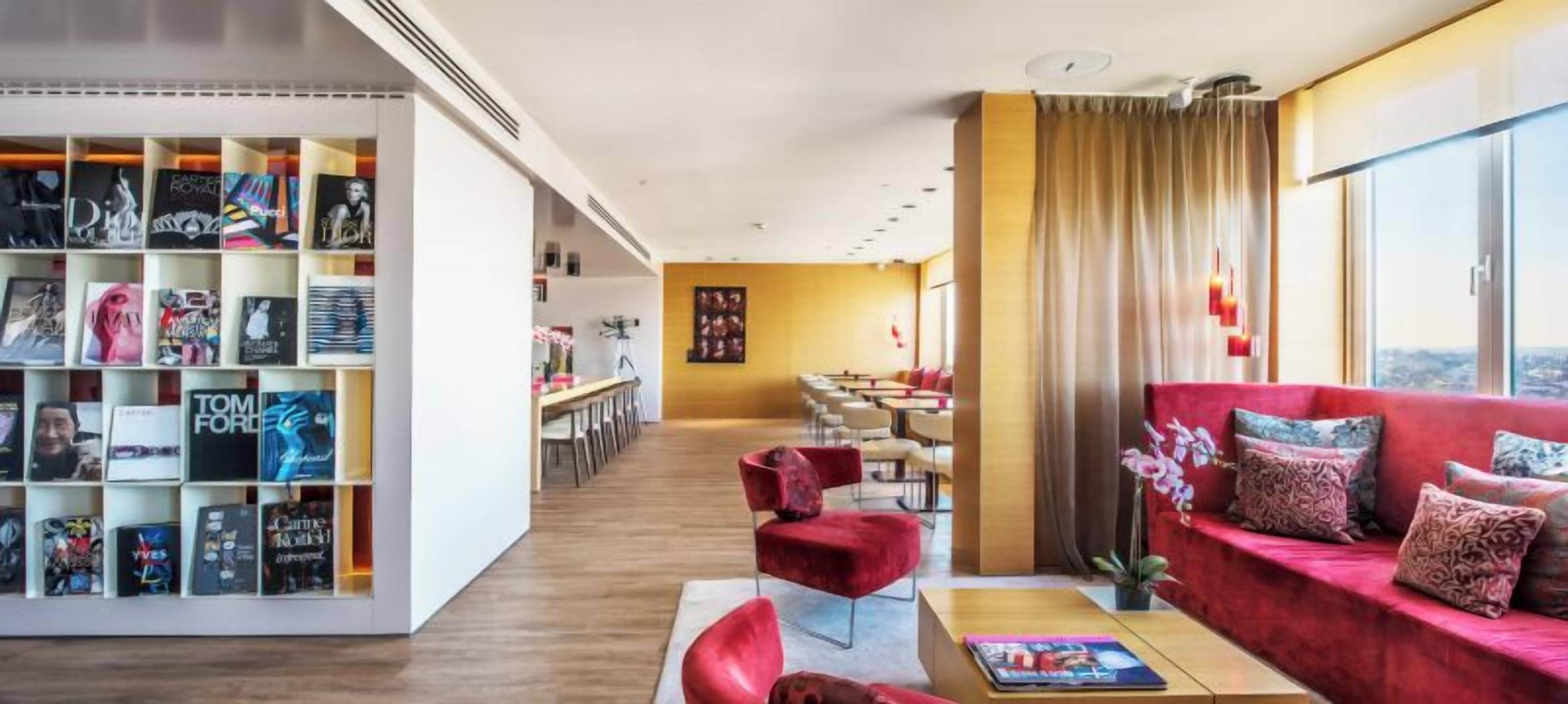 A modern hotel lobby with a red sofa, a bookshelf, and a dining area.